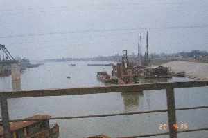 Thông tin văn bản duy nhất có thể đọc được là dấu thời gian kỹ thuật số ở góc dưới bên phải: '20 9:35'.。Bức ảnh ghi lại một công trường xây dựng quy mô lớn trên một con sông rộng. Khung cảnh được chụp từ một vị trí trên cao, có thể là từ trên một cây cầu hoặc một phương tiện đang di chuyển, với phần lan can kim loại ở tiền cảnh.。Dưới sông, nước có màu nâu đục, có nhiều phương tiện thủy đang hoạt động. Nổi bật là các sà lan (tàu) lớn và các giàn nổi được trang bị cần cẩu và máy móc xây dựng, cho thấy hoạt động thi công đang diễn ra. Có một chiếc thuyền nhỏ (ghe) đang di chuyển ở giữa sông.。Ở phía bên trái, có thể thấy một trụ cầu lớn bằng bê tông và một phần của kết cấu cầu giàn thép, gợi ý rằng đây là công trình xây dựng hoặc sửa chữa cầu. Dọc theo bờ sông phía xa bên phải là một bờ kè dốc được gia cố. Phía sau bờ kè là những ngôi nhà và cây cối. Bầu trời u ám và có các đường dây điện cao thế bắc ngang qua.。（日本語訳）。読み取れる唯一のテキスト情報は、右下隅にあるデジタルタイムスタンプ「20 9:35」です。この写真は、広い川で行われている大規模な建設現場を捉えています。川の水は茶色く濁っており、多くの水上車両が活動しています。目立っているのは、クレーンや建設機械を搭載した大型のはしけ（船）や浮き足場で、工事が行われていることを示しています。川の中央を小さなボート（ghe）が移動しています。左側には、大きなコンクリート製の橋脚と鋼製トラス橋の構造の一部が見え、これが橋の建設または修復工事であることを示唆しています。右奥の川岸に沿って、補強された傾斜のある護岸があります。護岸の後ろには家々や木々があります。空は曇っており、高圧電線が横切っています。