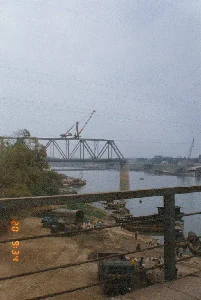 Bối cảnh là một công trường xây dựng bên một con sông lớn. Một cây cầu giàn thép đang trong quá trình thi công hoặc sửa chữa, với một chiếc cần cẩu đặt ở trên. Dưới sông, nhiều thuyền và sà lan đang neo đậu, có thể dùng để vận chuyển vật liệu xây dựng như cát, sỏi. Trên bờ sông bằng đất, có nhiều người đang làm việc, một số đội nón lá. Hoạt động của họ dường như là bốc dỡ vật liệu giữa thuyền và các xe tải đang đỗ gần đó. Có ít nhất hai chiếc xe tải có thể được nhìn thấy. Ở phía xa hơn trên sông cũng có một chiếc cần cẩu khác. Khung cảnh chung cho thấy một hoạt động xây dựng và vận tải tấp nập ven sông vào một ngày trời u ám. Ở góc dưới bên trái của hình ảnh có dấu thời gian màu cam ghi '20 9:34'.。（日本語訳）。背景は大きな川のほとりにある建設現場です。鉄骨トラス橋が建設中または修理中で、その上にクレーンが設置されています。川の下では、多くの船やはしけが停泊しており、砂や砂利などの建設資材を輸送するために使用されている可能性があります。土の川岸では、多くの人々が働いており、何人かはノンラーをかぶっています。彼らの活動は、船と近くに駐車しているトラックの間で資材を積み降ろししているようです。少なくとも2台のトラックが見えます。川のさらに遠くにも別のクレーンがあります。全体的な光景は、曇りの日に川沿いで行われる活発な建設および輸送活動を示しています。画像の左下隅には、オレンジ色のタイムスタンプで「20 9:34」と記されています。