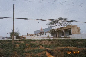 Bức ảnh chụp một tòa nhà bên đường vào năm 1994. Trên bức tường màu xanh của tòa nhà có dòng chữ lớn màu đỏ "KHU VĂN HÓA THỂ THAO" và dòng chữ nhỏ hơn màu đen bên dưới là "NHÀ MÁY DỆT VĨNH PHÚ". Đây là khu văn hóa thể thao của nhà máy dệt. Tòa nhà là một công trình một tầng, được bao quanh bởi hàng rào trắng. Phía trước là một bờ dốc đất có cỏ mọc. Khung cảnh còn có các cột điện, dây điện chằng chịt, và cây cối. Ở phía dưới, hình ảnh một người phụ nữ đi ngang qua bị mờ, cho thấy ảnh có thể được chụp khi đang di chuyển. Bầu trời xám xịt, phản ánh một khung cảnh đặc trưng của một thị trấn hoặc khu công nghiệp ở miền Bắc Việt Nam vào thời điểm đó.。（日本語訳）。1994年に撮影された、道端の建物の写真です。建物の青い壁には、赤い大きな文字で「KHU VĂN HÓA THỂ THAO」と書かれ、その下には黒い小さな文字で「NHÀ MÁY DỆT VĨNH PHÚ」と書かれています。これは紡績工場の文化・スポーツ地区です。建物は平屋建てで、白い柵で囲まれています。手前には草の生えた土手があります。下の方には、横切る女性の姿がぼやけており、移動中に撮影された可能性を示しています。どんよりとした空は、当時の北ベトナムの町や工業地帯の典型的な風景を反映しています。