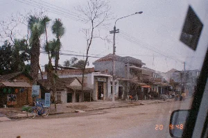 Bức ảnh chụp một con đường đất tại một thị trấn hoặc khu dân cư ở Việt Nam vào khoảng năm 1994. Khung cảnh được ghi lại từ bên trong một chiếc ô tô đang di chuyển, với một phần gương chiếu hậu của xe hiện rõ ở góc dưới bên phải.。**Thông tin văn bản:**。Ven đường có một số biển hiệu quảng cáo, tuy nhiên chữ viết đã mờ và khó đọc:。- Một tấm biển màu xanh lam dựng cạnh chiếc xe đạp, các chữ có thể đọc được là "SƠN SỈ" và "Ô TÔ".。- Một tấm biển khác gần đó có vẻ ghi "RỬA XE".。- Một tấm biển treo trên thân cây, chữ đã phai màu, dòng trên cùng có thể là "SỬA CHỮA".。- Trên cửa ra vào của một ngôi nhà hai tầng có một biển hiệu nhưng không thể đọc rõ nội dung.。**Mô tả cảnh vật và hoạt động:**。- **Loại địa điểm:** Đây là một con đường đất trong một khu dân cư hoặc thị trấn nhỏ. Hai bên đường là những ngôi nhà san sát nhau, bao gồm cả những ngôi nhà cấp bốn lợp ngói kiểu cũ và những ngôi nhà bê tông hai tầng, một số trông giống như cửa hàng hoặc cơ sở kinh doanh.。- **Hoạt động:** Khung cảnh trên đường khá yên tĩnh. Một người đang đứng ở cửa một ngôi nhà. Ở phía xa, có vài người đang di chuyển bằng xe đạp, trong đó có một người đội nón lá, một hình ảnh quen thuộc ở các vùng quê Việt Nam.。- **Các vật thể:**。- **Nhà cửa:** Dãy nhà phố với kiến trúc đa dạng, một số nhà còn đang xây dựng dở dang, phản ánh bối cảnh của thời kỳ đổi mới.。- **Phương tiện:** Một chiếc xe đạp màu xanh lam đang dựng bên lề đường. Phía xa có vài chiếc xe đạp và xe máy đang lưu thông.。- **Cơ sở hạ tầng:** Các cột điện, hệ thống dây điện chằng chịt và một cây đèn đường.。- **Thực vật:** Có những cây cọ và những cây thân gỗ khẳng khiu, trụi lá, cho thấy thời điểm chụp có thể là mùa đông hoặc đầu xuân ở miền Bắc.。（日本語訳）。1994年頃、ベトナムのある町または住宅地の未舗装の道を撮影した写真。風景は移動中の車内から記録されており、右下隅に車のサイドミラーの一部がはっきりと写っている。**テキスト情報：**。道端にはいくつかの広告看板があるが、文字はかすれていて読みにくい。- 自転車の隣に立てかけられた青い看板には、「SƠN SỈ」（卸売塗装）と「Ô TÔ」（自動車）という文字が読み取れる。- 近くにある別の看板には「RỬA XE」（洗車）と書かれているようだ。- 木の幹に掛けられた看板は文字が色褪せており、一番上の行は「SỬA CHỮA」（修理）かもしれない。- 2階建ての家の出入り口には看板があるが、内容ははっきりと読み取れない。**風景と活動の説明：**。- **場所の種類：** これは住宅地または小さな町の未舗装の道である。- **活動：** 道の上の風景はかなり静かである。一人の人が家の戸口に立っている。遠くには数人が自転車で移動しており、その中にはノンラー（菅笠）をかぶった人もいて、ベトナムの田舎ではおなじみの光景である。- **物体：**。- **家屋：** 多様な建築様式の町並みで、一部の家はまだ建設途中であり、ドイモイ（刷新）時代の背景を反映している。- **乗り物：** 青い自転車が道端に立てかけられている。遠くには数台の自転車やバイクが通行している。- **インフラ：** 電柱、複雑に絡み合った電線、そして街灯が一本ある。- **植物：** ヤシの木や、葉が落ちて痩せこけた木々があり、撮影時期が北部の冬または早春であることを示唆している。