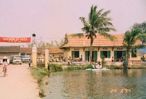 Bức ảnh chụp một con ngõ nhỏ và hẹp trong một ngôi làng ở vùng nông thôn, có lẽ là ở La Khê, tỉnh Hà Tây cũ. Lối đi được lát bằng gạch đỏ đã cũ. Bên trái là một phần của một ngôi nhà cấp bốn với tường vôi trắng và cửa sổ gỗ màu xanh lá cây. Bên phải là một bức tường dài, cũ kỹ, đã ngả màu và bám rêu mốc, trên đỉnh tường có thể có các mảnh chai để chống trộm. Dọc theo bức tường là cây cỏ mọc um tùm. Ở giữa lối đi có một thân cây cao, khẳng khiu, trông giống một cột điện bằng gỗ. Phía xa là một cổng vòm dẫn vào một khu vực khác trong làng. Có nhiều cây cối xanh tươi xung quanh, bao gồm cả cây đu đủ. Không có người hay hoạt động nào diễn ra. Khung cảnh mang vẻ yên bình, tĩnh lặng đặc trưng của làng quê Bắc Bộ Việt Nam thời kỳ đó.。（日本語訳）。写真は、田舎の村にある小さくて狭い路地を写しており、おそらく旧ハタイ省のラケーにある。通路は古い赤レンガで舗装されている。左側は、白い漆喰の壁と緑色の木製窓がある平屋建ての家の一部である。遠くには、村の別の区域に通じるアーチ型の門がある。周りにはパパイヤの木を含む、青々とした木々がたくさんある。人や活動は見られない。風景は、当時のベトナム北部の田舎に特有の、平和で静かな趣を帯びている。