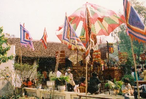 背景は1994年2月24日、北部農村地帯の広い空間、おそらくは村の集会所の庭か古い家の庭で行われている伝統的な村祭りです。イベントの中心は、朱漆と金箔の細工で精巧に装飾された練り歩き用の神輿です。神輿の上と周りには、大きな色とりどりの傘、竹の竿に立てられた五色の祭りの旗、大きな紙の扇子、そして「バッブウ」（八種の古代武器）のセットといった特徴的な儀式用の品々があります。神輿のあたりから線香の煙が立ち上っており、祭祀の儀式が行われていることを示しています。老人、大人、子供を含む非常に多くの地元の人々が、普段着を着て、祭りに参加し見物するために集まっています。何人かは壁のへりに座り、他の人々は立って見ています。ノンラー（菅笠）をかぶっている一人の女性がいます。後ろには、瓦屋根と白い壁を持つ伝統的な建築様式の家があります。家の壁には看板がありますが、書かれている文字は読むことができません。祭りの空間には、さらに木々や鉢植えの植物もあります。写真の右下隅には、「24 02 \'94」という黄色い日付の印があります