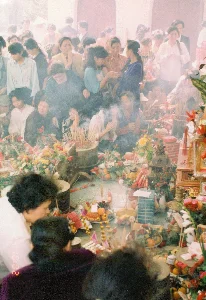 Bức ảnh ghi lại một khung cảnh đông đúc bên trong một không gian thờ cúng, có thể là một ngôi đình hoặc chùa, vào năm 1994. Không gian nghi ngút khói hương từ rất nhiều lư hương lớn đang cháy. Một đám đông người dân, chủ yếu là phụ nữ, đang thành kính tham gia một nghi lễ. Nhiều người đang chắp tay cầu nguyện (khấn vái), một số quỳ hoặc ngồi trên sàn. Trên sàn nhà và các ban thờ được bày biện rất nhiều lễ vật. Các mâm lễ vật bao gồm hoa tươi như hoa cúc, hoa hồng, các loại quả như cam, quýt, cùng với xôi và các vật phẩm vàng mã. Ở góc dưới bên trái bức ảnh có dấu thời gian kỹ thuật số màu đỏ ghi "'94 2 24 10:02". Trên các mâm lễ có những tờ giấy ghi chữ nhưng không thể đọc được nội dung.。（日本語訳）。写真は、1994年、ある礼拝空間、おそらくは đình（村の鎮守）か寺の内部の混雑した光景を記録しています。空間は、燃えている非常に多くの大きな香炉からの線香の煙が立ち込めています。主に女性からなる人々の群衆が、敬虔に儀式に参加しています。多くの人々が手を合わせて祈っており（拝んでおり）、一部の人は床にひざまずくか座っています。床の上と祭壇には、非常に多くの供物が供えられています。供物の盆には、菊やバラなどの生花、オレンジやみかんなどの果物類、おこわ、そして vàng mã（冥銭・紙製の供物）が含まれています。写真の左下隅には、「'94 2 24 10:02」と記された赤色のデジタルのタイムスタンプがあります。供物の盆の上には文字が書かれた紙がありますが、その内容を読むことはできません。