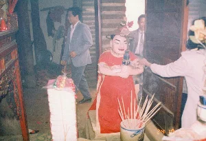 Bối cảnh bên trong một không gian thờ cúng, có thể là một ngôi đền hoặc đình làng, với không khí nghi lễ trang trọng. Ở trung tâm là một hình nhân lớn (ông phỗng) mặc áo choàng đỏ, đội mũ trang trí hoa, có khuôn mặt được tô vẽ đang mỉm cười. Một người phụ nữ đang đưa tay dâng tiền lễ cho hình nhân, các tờ tiền khác cũng được cài trên người hình nhân. Phía trước có một bát hương lớn bằng gốm sứ đang cắm đầy nhang cháy dở, khói tỏa nghi ngút. Bên trái là một phần của bàn thờ bằng gỗ sơn son được chạm khắc và một chồng lễ vật cao. Ở hậu cảnh, có hai người đàn ông mặc Âu phục đang có mặt trong không gian này. Ở góc dưới cùng bên phải của bức ảnh có dấu ngày tháng màu cam "24 10 94".。（日本語訳）。礼拝空間の内部の光景で、寺院か村の祠堂と思われ、荘厳な儀式の雰囲気が漂っている。中央には、赤い衣装をまとい、花で飾られた帽子をかぶり、微笑んでいるように描かれた顔を持つ大きな人形（オン・フォッン）がある。一人の女性が人形にお布施を捧げようと手を伸ばしており、他の紙幣も人形の体に差し込まれている。手前には、燃えかけの線香がたくさん立てられた大きな陶磁器製の香炉があり、煙がもうもうと立ち上っている。左側には、彫刻が施された朱塗りの木製祭壇の一部と、高く積まれたお供え物がある。背景には、洋服を着た二人の男性がこの空間にいる。写真の右下隅には、「24 10 94」というオレンジ色の日付印がある。