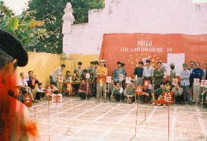 Trên tấm phông lớn màu đỏ có dòng chữ: 'HỘI CỜ LÀNG LA KHÊ XUÂN GIÁP TUẤT 1994'. Trên các tấm biển dùng làm quân cờ có các chữ Hán như: 車 (Xe), 卒 (Tốt), 士 (Sĩ), 象 (Tượng), 炮 (Pháo).。Bối cảnh là một sân rộng, có thể là sân đình hoặc không gian sinh hoạt chung tại làng La Khê, tỉnh Hà Tây. Một lễ hội cờ người đang được tổ chức, đây là một hoạt động văn hóa truyền thống trong dịp lễ hội mùa xuân. Rất đông người dân, bao gồm cả đàn ông và trẻ em, đang tụ tập để xem thi đấu. Trên sân đất được kẻ các ô vuông lớn mô phỏng bàn cờ tướng. Một số người đóng vai các quân cờ, đứng trên bàn cờ và cầm các tấm biển ghi tên quân cờ bằng chữ Hán. Khán giả ngồi và đứng xung quanh để theo dõi ván đấu trong không khí sôi nổi. Phía sau có những bức tường cũ màu vàng và trắng cùng với lùm cây chuối.。（日本語訳）。大きな赤い背景幕には、「HỘI CỜ LÀNG LA KHÊ XUÂN GIÁP TUẤT 1994」（ラケー村の人間将棋祭り 甲戌の春 1994年）という文字が書かれている。駒として使われる札には、車（セ）、卒（トット）、士（シー）、象（トゥオン）、炮（ファオ）などの漢字がある。背景は広い庭で、ハタイ省ラケー村の村の集会所の庭か、共同生活空間である可能性がある。人間将棋の祭りが開催されており、これは春の祭りにおける伝統的な文化活動である。男性や子供を含む非常に多くの村人が、試合を観戦するために集まっている。土の地面には、シャンチー（中国将棋）の盤を模した大きな四角いマスが引かれている。何人かの人々が駒の役を演じ、盤の上に立ち、漢字で駒の名前が書かれた札を持っている。観客は周りに座ったり立ったりして、活気あふれる雰囲気の中で対局を見守っている。後方には、黄色と白の古い壁とバナナの茂みがある。