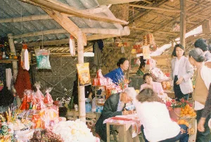 Bối cảnh là một gian hàng trong một khu chợ ở vùng quê, có kết cấu đơn sơ với mái lợp tôn và lá, cột và tường làm bằng tre, gỗ. Không khí mua bán khá nhộn nhịp với nhiều người có mặt.。Hoạt động chính diễn ra tại gian hàng là mua bán các mặt hàng, chủ yếu là đồ lễ cúng. Một cụ ông tóc bạc, mặc áo vest, đang ngồi tại một chiếc bàn gỗ nhỏ và cặm cụi dùng bút viết lên giấy, có thể là một thầy đồ đang viết chữ để bán. Một người phụ nữ mặc áo xanh, có lẽ là chủ hàng, đang đứng sau quầy. Xung quanh có nhiều khách hàng, chủ yếu là phụ nữ, đang lựa chọn hàng hóa.。Các vật thể có trong ảnh bao gồm: nhiều phẩm oản hình tháp được gói giấy bóng kính màu đỏ, bánh kẹo, hoa quả (nho, quýt), hoa tươi cắm trong xô, và hương (nhang). Ở phía sau quầy có một chiếc cân đĩa màu xanh. Nhiều gói hàng được treo trên các xà nhà bằng tre. Một số bao bì sản phẩm có chữ nhưng không thể đọc rõ nội dung.。（日本語訳）。背景は田舎の市場にある一軒の露店で、トタンと葉で葺かれた屋根、竹と木で作られた柱と壁という簡素な造りである。売買の雰囲気はかなり賑やかで、多くの人がいる。露店で行われている主な活動は品物の売買であり、主にお供え物である。白髪の老人男性が一人、ベストを着て、小さな木製の机に座り、熱心にペンで紙に何かを書いている。おそらく販売用の文字を書いている書道の先生だろう。青い服を着た女性が一人、おそらく店主で、カウンターの後ろに立っている。周りには多くの客がおり、主に女性で、商品を選んでいる。写真に写っている物には、赤いセロハンで包まれた塔の形をした多くのお供え餅（オアン）、菓子、果物（ブドウ、ミカン）、バケツに生けられた生花、そして線香が含まれる。カウンターの後ろには、青い皿秤が一つある。多くの包みが竹でできた梁に吊るされている。いくつかの商品のパッケージには文字があるが、内容をはっきりと読み取ることはできない。