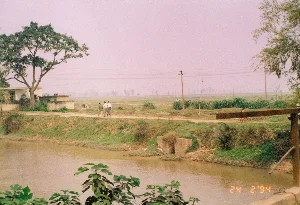 Bức ảnh chụp một khung cảnh nông thôn yên bình, có khả năng là ở khu vực La Khê, tỉnh Hà Tây vào ngày 24 tháng 2 năm 1994. Ở phía trước là một con sông hoặc con kênh với dòng nước màu nâu. Dọc theo bờ sông là một con đường đất, nơi có ba người đàn ông đang đi bộ và trò chuyện. Bên trái có một cây cổ thụ lớn và một vài ngôi nhà cấp 4 đơn sơ. Phía sau con đường là những cánh đồng nông nghiệp trải dài ra xa, với các cột điện và đường dây điện chạy ngang qua. Ở phía xa trên con đường, có thể thấy một người đang đi xe đạp màu đỏ. Bầu trời có màu hồng nhạt, có thể do thời tiết hoặc thời điểm chụp. Dưới góc phải của ảnh có dấu ngày tháng được ghi là '24 2 '94'. Không có văn bản nào khác như biển báo hay chữ khắc.。（日本語訳）。この写真は、1994年2月24日に、おそらくハタイ省ラケー地区で撮影された、平和な田園風景を写したものです。手前には、茶色の水が流れる川か水路があります。川岸に沿って土の道があり、そこを3人の男性が歩きながら話しています。左側には、大きな古木が1本と、いくつかの簡素な平屋があります。道の後ろには農地が遠くまで広がっており、電柱と電線が横切っています。道の遠くの方には、赤い自転車に乗っている人が一人見えます。空は薄いピンク色をしており、天候または撮影時間によるものかもしれません。写真の右下隅には、「'24 2 '94」と記された日付スタンプがあります。標識や刻印のような他のテキストはありません。