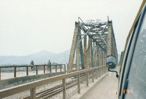 Bức ảnh được chụp từ một phương tiện đang di chuyển, có lẽ là ô tô, khi đi qua một cây cầu giàn thép lớn. Dựa vào bối cảnh địa lý (Thanh Hóa) và kiến trúc, đây rất có thể là Cầu Hàm Rồng lịch sử. Trên đỉnh của giàn thép có dòng chữ số "1965". Cây cầu này có kết cấu phức hợp bao gồm cả đường bộ và đường sắt chạy song song.。Trên làn đường dành cho xe thô sơ của cây cầu, có hai người đang đi xe đạp. Một người trong số họ đội nón lá. Hoạt động chính là giao thông qua cầu.。Các vật thể có trong ảnh bao gồm: cây cầu giàn thép, đường ray xe lửa, hai chiếc xe đạp, và một phần của chiếc ô tô (bao gồm gương chiếu hậu) mà từ đó bức ảnh được chụp. Phía sau là khung cảnh đồi núi lờ mờ dưới bầu trời có mây. Dấu ngày tháng "26 3 '94" được in trên ảnh.。（日本語訳）。写真は、移動中の乗り物、おそらくは自動車から、大きな鋼鉄のトラス橋を渡る際に撮影されたものです。地理的な背景（タインホア省）と建築様式から、これは歴史的なハムロン橋である可能性が非常に高いです。鋼鉄トラスの上部には「1965」という数字があります。この橋は、道路と鉄道が並行して走る複合的な構造をしています。橋の軽車両用の車線には、自転車に乗っている人が2人います。そのうちの一人はノンラーをかぶっています。主な活動は橋を渡る交通です。写真に写っている物体には、鋼鉄のトラス橋、線路、2台の自転車、そして写真が撮影された自動車の一部（バックミラーを含む）が含まれます。後方には、曇り空の下にぼんやりとした丘や山の風景が広がっています。写真には「26 3 '94」という日付が印字されています。