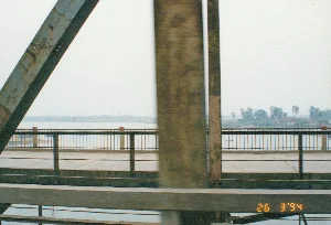 Bức ảnh được chụp từ một phương tiện đang di chuyển khi đi qua một cây cầu lớn bằng thép, có thể là ở khu vực Thanh Hóa. Trên ảnh có dấu ngày tháng màu vàng "26 3 '94". Các kết cấu dầm thép của cây cầu chiếm phần lớn tiền cảnh và bị mờ do chuyển động. Bên dưới là một con sông rộng lớn. Phía xa bên kia sông là bờ đất với cây cối và một vài công trình nhà cửa thấp tầng. Có một chiếc thuyền nhỏ đang ở trên sông. Toàn bộ khung cảnh chìm trong một lớp sương mù mờ ảo.。（日本語訳）。この写真は、おそらくタインホア省の地域で、大きな鋼鉄の橋を渡る際に移動中の乗り物から撮影されたものです。写真には「94年3月26日」という黄色い日付スタンプがあります。橋の鋼鉄の梁の構造が前景の大部分を占めており、動きによってぼやけています。下には広大な川があります。川の向こう岸の遠くには、木々やいくつかの低層の建物がある土手があります。川の上には小さな船が一艘あります。風景全体が、ぼんやりとした霧の層に包まれています。