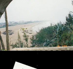 Góc dưới bên phải của hình ảnh có dấu thời gian kỹ thuật số màu cam ghi: "26 13:32 92".。Đây là một bức ảnh chụp toàn cảnh từ trên cao, cho thấy một khu dân cư ở một thị trấn hoặc làng quê tại Việt Nam, có thể là ở khu vực Thanh Hóa, vào năm 1994. Khung cảnh chủ yếu là các mái nhà lợp ngói san sát nhau. Các ngôi nhà có kiến trúc đa dạng, một số là nhà gạch mộc cũ kỹ, trong khi những ngôi nhà khác được trát vữa và sơn màu (vàng, trắng). Mái nhà chủ yếu là mái ngói đất nung màu nâu sẫm hoặc đỏ, một đặc trưng của kiến trúc nhà ở truyền thống Việt Nam. Trên một số mái nhà có lắp đặt ăng-ten TV, một vật dụng phổ biến vào thời điểm đó. Xen kẽ giữa các ngôi nhà là cây cối xanh tươi. Ở phía trước, nổi bật là những bụi chuối lớn với tán lá xanh mướt. Bầu trời có nhiều mây và không có hoạt động nào của con người được nhìn thấy, tạo nên một không gian yên tĩnh.。（日本語訳）。画像の右下隅には、「26 13:32 92」と記されたオレンジ色のデジタルタイムスタンプがあります。これは、1994年にベトナムのある町または田舎、おそらくタインホア地方の住宅地を上から撮影した全景写真です。主な風景は、密集した瓦葺きの屋根です。家々は多様な建築様式で、いくつかは古いレンガむき出しの家である一方、他の家は漆喰が塗られ、（黄色、白の）色で塗装されています。屋根は主に濃い茶色または赤色の素焼きの瓦で、ベトナムの伝統的な住宅建築の特徴です。いくつかの屋根の上には、当時一般的だったテレビアンテナが設置されています。家々の間には、青々とした木々が点在しています。手前には、青々とした葉を持つ大きなバナナの茂みが目立っています。空は雲が多く、人の活動は見られず、静かな空間を作り出しています。