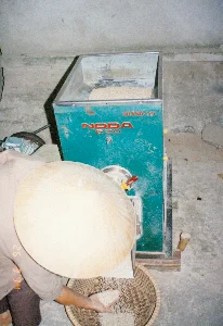 緑色の精米機の本体には、「NODA 620」と「THÀNH LỢI」という赤い文字がある。背景は小さな精米所で、作業場か、コンクリートの床がある農村地域の民家の一角かもしれない。農民と思われる、ノンラー（菅笠）をかぶった人が機械を操作している。籾（もみ、未精米の米）は機械の上部にあるホッパーに入れられている。精米された白米が機械から、下に置かれた竹で編んだ大きな平かごに流れ落ちている。この人は身をかがめ、手のひらで精米したての米粒を受け止め、品質を確認している