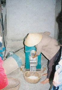 Trong ảnh có dấu ngày tháng màu đỏ "26 3 '94". Bối cảnh là một không gian trong nhà, có vẻ là một cơ sở xay xát lúa gạo nhỏ ở vùng nông thôn. Một người đang đội nón lá, mặc áo khoác màu nâu và đi chân đất, đang vận hành một chiếc máy xay xát lúa màu xanh. Người này cúi xuống, dùng tay để điều chỉnh hoặc kiểm tra dòng gạo trắng đang chảy từ máy ra. Gạo sau khi xay được hứng vào một chiếc rổ tre đan đặt ngay dưới cửa ra của máy. Bên cạnh đó còn có một chiếc rổ tre khác. Không gian xung quanh khá đơn sơ với sàn và tường bằng xi măng.。（日本語訳）。写真には「94年3月26日」という赤い日付スタンプがあります。背景は屋内の空間で、田舎の小さな精米所のようです。ノンラー（菅笠）をかぶり、茶色の上着を着て裸足の人が、青い精米機を操作しています。この人はかがみ込み、手を使って機械から流れ出る白米の流れを調整または確認しています。精米された米は、機械の排出口の真下に置かれた竹で編んだかごに受けられています。その隣には、もう一つ別の竹かごがあります。周囲の空間は、床と壁がセメントでできており、かなり質素です。