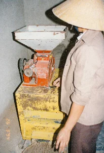 Trong ảnh có dấu ngày tháng '26 3 '94'. Bức ảnh ghi lại cảnh một người phụ nữ đội nón lá đang làm việc bên một chiếc máy xay xát. Khung cảnh diễn ra trong một không gian trong nhà, có thể là một cơ sở chế biến nông sản nhỏ hoặc nhà kho với những bức tường xi măng. Người phụ nữ mặc áo khoác màu be, đang đứng cạnh chiếc máy để vận hành hoặc giám sát. Chiếc máy có phần thân màu đỏ và bệ đỡ màu vàng, phía trên là một cái phễu lớn màu trắng để chứa nguyên liệu. Dường như máy đang hoạt động, sản phẩm sau khi xay (có thể là cám hoặc thức ăn gia súc dạng viên) đang chảy từ máy xuống một chiếc rổ hoặc thúng đặt trên sàn nhà.。（日本語訳）。写真には「'94 3 '26」という日付スタンプがあります。この写真は、ノンラーをかぶった女性が精米機のそばで作業している光景を記録しています。その光景は屋内で繰り広げられており、セメントの壁がある小さな農産物加工施設か倉庫かもしれません。女性はベージュの上着を着て、機械を操作または監視するためにその隣に立っています。機械は本体が赤色で土台が黄色、上部には原料を入れるための大きな白い漏斗があります。機械は作動しているようで、粉砕後の製品（米ぬかまたはペレット状の家畜飼料である可能性があります）が機械から床に置かれたかごまたはバスケットに流れ落ちています。