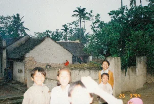 Trên ảnh có dấu ngày tháng màu vàng "26 3 '94". Bức ảnh chụp lại một khoảnh khắc đời thường tại một làng quê ở Việt Nam, có thể là ở khu vực Thanh Hóa, vào năm 1994. Trong ảnh là một nhóm trẻ em đang vui vẻ tạo dáng trước ống kính. Bối cảnh chính là một ngôi nhà cấp 4 truyền thống với tường gạch trát vữa đã cũ và bong tróc ở một vài chỗ, để lộ ra lớp gạch đỏ bên trong. Mái nhà được lợp bằng ngói ta, đã sẫm màu vì thời gian. Xung quanh và phía sau ngôi nhà là cây cối xanh tươi, rậm rạp, có thể thấy những cây cao như cây dừa và cây cau. Ở tiền cảnh và trung tâm bức ảnh, một nhóm khoảng 5-6 trẻ em đang tươi cười. Một số em đứng gần một bức tường bê tông thấp. Trang phục của các em khá đơn giản, phản ánh cuộc sống thời kỳ đó. Một cánh tay và phần đầu của một người bị quá sáng ở phía trước che mất một phần khung cảnh. Ở phía sau, gần ngôi nhà, cũng có thể thấy một vài đứa trẻ khác.。（日本語訳）。写真には「94年3月26日」という黄色い日付スタンプがある。この写真は、1994年にベトナムの田舎、おそらくタインホア地方での日常の一コマを捉えたものである。写真には、カメラの前で楽しそうにポーズをとる子供たちのグループが写っている。主な背景は、伝統的な平屋建ての家で、漆喰塗りのレンガ壁は古くなり、所々剥がれて中の赤いレンガが露出している。屋根はベトナム瓦で葺かれており、時を経て黒ずんでいる。写真の前景と中央には、5〜6人ほどの子供たちのグループが笑顔で写っている。何人かの子供は低いコンクリートの壁の近くに立っている。子供たちの服装は非常にシンプルで、当時の生活を反映している。前景で露出オーバーになった人の腕と頭の一部が、景色の一部を覆っている。