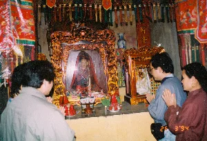 Bức ảnh chụp cảnh bên trong một ngôi chùa, đền hoặc điện thờ vào ngày 26 tháng 3 năm 1994. Có ba người, gồm hai người đàn ông và một người phụ nữ, đang đứng trước một bàn thờ lớn và thực hiện nghi lễ cúng bái. Họ chắp hai tay trước ngực trong tư thế cầu nguyện thành kính. Bàn thờ được chạm khắc công phu, sơn son thếp vàng. Ở trung tâm là một khám thờ lớn, bên trong có tượng hoặc ảnh thờ một vị thần được phủ khăn đỏ. Trên bàn thờ bày các vật phẩm cúng lễ như bát nhỏ, nến đang cháy và các tháp oản phẩm hoặc giấy tiền vàng mã màu đỏ. Không gian được trang trí bằng các tấm trướng, cờ phướn nhiều màu sắc có thêu hình rồng và hoa văn truyền thống. Phía sau bàn thờ có một bức hoành phi bằng gỗ màu đỏ khắc chữ Hán/Nôm theo chiều dọc, nhưng các ký tự khó đọc rõ, và một chiếc lọ hoa bằng gốm sứ men lam trắng.。（日本語訳）。1994年3月26日に撮影された、ある寺院、祠、または宮殿の内部の光景を写した写真です。男性2人と女性1人の計3人が、大きな祭壇の前に立ち、礼拝の儀式を行っています。彼らは敬虔な祈りの姿勢で胸の前で両手を合わせています。祭壇は精巧に彫刻され、朱塗り金箔で装飾されています。中央には大きな祠があり、その中には赤い布で覆われた神様の像または肖像画が祀られています。祭壇の上には、小さな碗、燃えているろうそく、そしてお供え物や赤い冥銭の塔などが供えられています。空間は、龍の刺繍や伝統的な文様が施された色とりどりの垂れ幕や幟で飾られています。祭壇の後ろには、縦書きで漢字/チュノムが刻まれた赤い木製の扁額がありますが、文字ははっきりと読み取るのが難しく、また青と白の染付の陶磁器の花瓶があります。