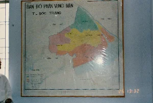 Bối cảnh là một căn phòng, có thể là văn phòng hoặc phòng họp. Trên bức tường màu xanh nhạt có treo một tấm bản đồ lớn trong khung gỗ, được bọc một lớp nhựa trong suốt gây phản quang.。Chữ viết trên bản đồ:。- Tiêu đề chính: "BẢN ĐỒ PHÂN VÙNG MẶN T_ SÓC TRĂNG".。- Tên các tỉnh lân cận: "T. CẦN THƠ", "T. TRÀ VINH", "T. MINH HẢI".。- Tên các huyện, thị xã trong tỉnh Sóc Trăng: "KẾ SÁCH", "MỸ TÚ", "THẠNH TRỊ", "TX SÓC TRĂNG", "MỸ XUYÊN", "LONG PHÚ".。- Tiêu đề bảng chú giải: "CHÚ DẪN". Nội dung chi tiết trong bảng không thể đọc rõ.。- Có một dòng chữ ở cuối bản đồ nhưng bị mờ và khó đọc.。Hoạt động và vật thể:。- Địa điểm: Văn phòng hoặc phòng họp.。- Hoạt động: Trưng bày bản đồ để tham khảo hoặc phục vụ công việc. Đây là bản đồ kỹ thuật về độ nhiễm mặn của đất và nước, cho thấy công việc có thể liên quan đến nông nghiệp, thủy lợi hoặc quản lý tài nguyên môi trường tại tỉnh Sóc Trăng.。- Vật thể: Tấm bản đồ phân vùng nhiễm mặn tỉnh Sóc Trăng, khung gỗ, tường, một phần cửa sổ ở bên trái và một phần hình ảnh của một người mặc áo sơ mi trắng.。（日本語訳）。背景は部屋で、オフィスまたは会議室かもしれません。淡い青色の壁には、木製の額縁に入った大きな地図が掛けられており、光を反射する透明なプラスチックで覆われています。地図上の文字：。- メインタイトル：「BẢN ĐỒ PHÂN VÙNG MẶN T_ SÓC TRĂNG」。- 近隣の省名：「T. CẦN THƠ」、「T. TRÀ VINH」、「T. MINH HẢI」。- ソクチャン省内の県、町の名前：「KẾ SÁCH」、「MỸ TÚ」、「THẠNH TRỊ」、「TX SÓC TRĂNG」、「MỸ XUYÊN」、「LONG PHÚ」。- 凡例のタイトル：「CHÚ DẪN」。表内の詳細な内容ははっきりと読み取れません。- 地図の下部に一行の文字がありますが、ぼやけていて読みにくいです。活動と物体：。- 場所：オフィスまたは会議室。- 活動：参考または業務のために地図を展示。これは土壌と水の塩害度に関する技術的な地図であり、業務がソクチャン省における農業、水利、または環境資源管理に関連している可能性を示しています。- 物体：ソクチャン省の塩害区分図、木製の額縁、壁、左側にある窓の一部、そして白いシャツを着た人物の姿の一部。