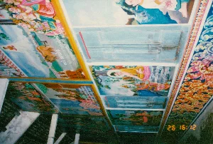 Bức ảnh chụp trần nhà của một công trình kiến trúc, rất có thể là một ngôi chùa Phật giáo Khmer ở Sóc Trăng. Toàn bộ trần nhà được chia thành nhiều ô vuông và được trang trí bằng các bức tranh tường sặc sỡ. Các bức tranh này mô tả những cảnh trong Phật giáo, có thể là về cuộc đời của Đức Phật hoặc các câu chuyện tiền thân (Jataka). Trong tranh có hình ảnh Đức Phật, các vị chư thiên, con người trong trang phục truyền thống và động vật như voi. Bên dưới một số bức tranh có các dòng chữ, dường như là chữ Khmer, nhưng không thể đọc rõ nội dung. Viền trần nhà được trang trí bằng những hoa văn truyền thống rất công phu và nhiều màu sắc. Một phần kết cấu của tòa nhà với các cột và dầm bê tông cũng có thể được nhìn thấy ở phía dưới bên trái.。（日本語訳）。ある建築物の天井を撮影した写真で、おそらくソクチャン省にあるクメール仏教寺院のものでしょう。天井全体は多くの四角い区画に分けられ、色鮮やかな壁画で装飾されています。これらの絵は仏教の場面を描写しており、仏陀の生涯や本生話（ジャータカ）に関するものである可能性があります。絵の中には、仏陀、天人、伝統的な衣装をまとった人々、そして象のような動物の姿があります。いくつかの絵の下には文字の列があり、クメール文字のようですが、内容ははっきりと読み取れません。天井の縁は、非常に精巧で色彩豊かな伝統的な文様で装飾されています。左下には、コンクリートの柱や梁といった建物の構造の一部も見ることができます。