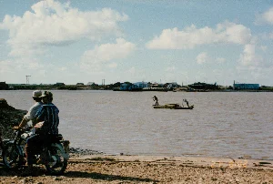 Không có thông tin dạng chữ viết nào có thể đọc được trong ảnh.。Bức ảnh ghi lại cảnh một nhóm đông người, bao gồm đàn ông, phụ nữ và trẻ em, đang tụ tập tại một khu vực nuôi trồng thủy sản ở vùng nông thôn, có thể là ở Sóc Trăng. Địa điểm là một khu đất với các ao, mương nước được đào để nuôi tôm hoặc cá. Hoạt động chính đang diễn ra là thu hoạch thủy sản. Một số người đang đứng dưới ao, kéo các lồng lưới màu xanh để bắt sản phẩm. Những người khác đứng trên bờ đất để quan sát hoặc phụ giúp. Khung cảnh cho thấy một buổi lao động tập thể. Các vật thể có trong ảnh bao gồm: nhiều người dân mặc trang phục lao động đơn giản, một số người đội nón lá, các lồng lưới nuôi thủy sản, một túp lều lợp lá đơn sơ ở phía xa, và các dụng cụ như một cái cân và một cái thau nhựa được đặt trên bờ. Xung quanh là cây cối rậm rạp, đặc trưng của vùng sông nước.。（日本語訳）。写真には読み取れる文字情報はありません。この写真は、男性、女性、子供を含む大勢の人々が、おそらくソクチャン省の田舎にある水産養殖場で集まっている場面を記録しています。場所は、エビや魚を養殖するために掘られた池や水路のある土地です。行われている主な活動は水産物の収穫です。何人かの人々は池の中に立ち、青い網かごを引いて収穫物を捕っています。他の人々は土手の上に立って見物したり手伝ったりしています。この光景は共同作業の様子を示しています。写真に写っている物には、簡素な作業着を着た多くの人々、ノンラーをかぶった数人、水産養殖用の網かご、遠くにある簡素な葉葺きの小屋、そして土手の上に置かれた秤やプラスチック製のたらいのような道具が含まれます。周囲は、水郷地帯に特徴的な、生い茂った木々です。