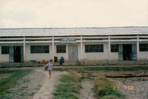 1994年に撮影された、農村地帯にある小学校の写真です。建物の正面に取り付けられた青い看板には「タイン・トーイ・トゥアン第二小学校」と書かれています。これは平屋建て、1階建て、セメント瓦屋根の建物で、鉄格子の窓と青く塗られた出入り口のある教室が複数あります。学校の前は未舗装の道と、収穫後の田んぼかもしれない空き地です。ストライプのシャツを着た少年が未舗装の道を歩いています。校舎の軒先では、数人が座って休んでいます。近くに自転車が一台停められています。全体的な光景は、ある田舎ののどかな朝を示しています