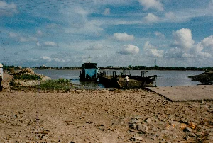 Bối cảnh là một bến phà ven sông ở một vùng nông thôn vào năm 1994. Mặt đất ở bờ sông là đất bùn và đá lởm chởm. Một chiếc phà có cabin màu xanh đang cập vào một cầu tàu bằng bê tông đơn sơ. Trên phà, một người đàn ông mặc áo trắng đang đứng. Ở phía xa bên kia sông, có thể thấy một vài ngôi nhà và cây cối. Một cột điện cao thế cũng hiện ra ở phía xa. Bầu trời xanh và có nhiều mây trắng. Ở bên trái bức ảnh, có một người đang đội nón lá. Hoạt động chính là chiếc phà đang chuẩn bị hoặc vừa hoàn thành việc vận chuyển người và có thể cả phương tiện qua sông, một hình thức giao thông phổ biến ở miền Tây Nam Bộ thời kỳ này. Ở góc dưới bên phải ảnh có dấu ngày tháng màu cam ghi '27 8:16'.。（日本語訳）。舞台は1994年、ある田舎の川沿いの渡し場です。川岸の地面は泥とごつごつした石です。青い船室のある一隻のフェリーが、簡素なコンクリートの桟橋に接岸しています。フェリーの上には、白いシャツを着た一人の男性が立っています。川の向こう岸の遠くには、数軒の家と木々が見えます。遠くには高圧電線の鉄塔も見えます。空は青く、白い雲がたくさん浮かんでいます。写真の左側には、ノンラーをかぶった人が一人います。主な活動は、フェリーが川を渡る人や、場合によっては車両の輸送を準備しているか、あるいは終えたところであり、これはこの時代のメコンデルタ地域で一般的な交通手段です。写真の右下隅には、「27 8:16」と書かれたオレンジ色の日付スタンプがあります。