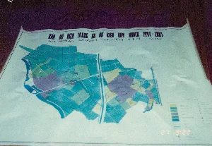 Bức ảnh chụp cận cảnh một tấm bản đồ quy hoạch bằng giấy được trải trên một bề mặt tối màu, có thể là một chiếc bàn. Các mép của tờ giấy hơi cong lên. Đây là bản đồ thể hiện hiện trạng và kế hoạch sử dụng đất của một khu vực, được phân chia thành nhiều lô đất với các màu sắc khác nhau như xanh lam, xanh lá, tím và vàng. Các đường kẻ trên bản đồ có thể là hệ thống sông ngòi, kênh rạch hoặc đường giao thông.。Nội dung chữ viết trên bản đồ được ghi lại như sau:。- Tiêu đề chính: "BẢN ĐỒ HIỆN TRẠNG VÀ DỰ KIẾN QUY HOẠCH 1993-2005"。- Địa danh: "XÃ ĐỒNG QUANG HUYỆN TIÊN SƠN"。- Tỷ lệ: "TỶ LỆ 1:5000" (chữ số hơi mờ)。- Tiêu đề phần chú thích: "CHÚ DẪN" (nội dung chi tiết không đọc được).。（日本語訳）。暗い色の表面、おそらくテーブルの上に広げられた紙の計画地図のクローズアップ写真です。紙の端は少し上に反っています。これは、ある地域の土地利用の現状と計画を示す地図で、青、緑、紫、黄色など様々な色で多くの区画に分けられています。地図上の線は、河川や運河のシステム、または交通路である可能性があります。地図に書かれている文字の内容は以下の通りです。- メインタイトル：「1993-2005年 現状および計画予測図」。- 地名：「ティエンソン県ドンクアン社」。- 縮尺：「縮尺 1:5000」（数字はやや不鮮明）。- 凡例のタイトル：「凡例」（詳細な内容は読み取れない）。