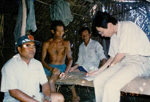 場面は、ソクチャン省のある農村地域と思われる、簡素な葉葺きの家の中で展開されている。家の屋根と壁はニッパヤシの葉で葺かれており、構造は簡素な木の柱で支えられている。写真には4人の男性が写っている。3人の男性は地元住民のように見え、もう1人の男性は眼鏡をかけ、明るい色のシャツとズボンを着用しており、インタビューか調査を行っている研究者または職員のようだ。この職員は竹製の寝台に座り、ペンで紙に熱心に何かを書き留めている。3人の住民のうち、1人の男性はカメラの方を向いて座り、野球帽（帽子には赤い文字があるがはっきりと読めない）をかぶり、シャツを着ている。竹製の寝台で彼の隣に座っているのは、痩せた年配の男性で、上半身は裸で青い半ズボンを履いている。3人目の人物は彼らの後ろに座っている。全員、ござが敷かれた竹製の寝台の上に座っている。上の方、家の梁には数着の服が掛かっている。写真の右下隅には、「27 15:51」と記されたオレンジ色のデジタルのタイムスタンプがある。この光景は、1994年に人々の質素な家の中で行われた会合、情報交換の様子を示している