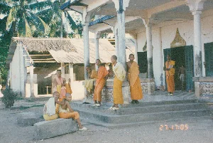 この写真は、若い僧侶たち、おそらくは見習い僧の一団が、ある寺の軒先で休憩している光景を捉えている。背景は農村地帯のようであり、地理的な背景からソクチャン地域のクメール寺院である可能性があり、それと一致している。僧侶たちは頭を剃髪し、オレンジ色と黄色の袈裟を身に着けている。彼らはくつろいだ様子で集まっている。数人はセメントの階段に座り、他の者たちは軒の柱に寄りかかって立ち、会話をしながら笑顔でカメラの方を見ている。寺は簡素な建築様式で、軒と丸いコンクリートの柱があり、白い壁は古びて一部が剥がれ落ちている。出入り口の扉は緑色の木製ルーバー扉で、その上には伝統的な様式の彫刻装飾が施されている。周囲には土の庭と高いヤシの木々が並び、メコンデルタ地域特有の平穏な風景を作り出している。写真の右下隅には、「27 17:05」と記されたオレンジ色のデジタルタイムスタンプがある。他に認識できるテキストはない