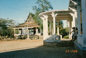 Bối cảnh là khuôn viên của một ngôi chùa Khmer ở Sóc Trăng. Ở phía sau là một công trình chùa với mái ngói nhiều tầng có các chi tiết trang trí cong vút đặc trưng của kiến trúc Khmer. Phần chân tường rào của công trình này được trang trí bằng các họa tiết hình bánh xe, có thể là Bánh xe Pháp (Dharmachakra). Phía trước, nơi chụp ảnh, là một dãy hành lang hoặc nhà sala màu trắng, có mái che và nhiều cột trụ. Sân chùa là một khoảng sân đất rộng. Ở bên phải bức ảnh, một vài cậu bé đang ngồi trên bậc thềm của dãy hành lang. Có một cột cờ cao được dựng giữa sân. Dấu thời gian kỹ thuật số ở góc dưới bên phải ghi '27 17:06'.。（日本語訳）。背景はソクチャン省にあるクメール寺院の境内です。後方には、クメール建築に特徴的な、反り上がった装飾的ディテールを持つ多層の瓦屋根の寺院建築があります。この建物の塀の土台部分は、法輪（ダルマチャクラ）かもしれない車輪の形のモチーフで装飾されています。手前、撮影場所には、屋根と多くの柱がある白い回廊またはサラ（sala）の列があります。寺の境内は広い土の庭です。写真の右側では、数人の少年が回廊の階段に座っています。庭の中央には高い旗竿が立てられています。右下のデジタルタイムスタンプには「27 17:06」と記されています。