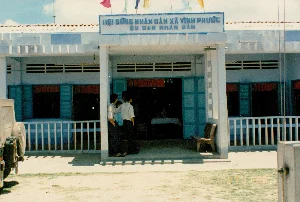 Trên biển hiệu màu xanh lam phía trên lối vào tòa nhà có dòng chữ trắng: 'HỘI ĐỒNG NHÂN DÂN XÃ VĨNH PHƯỚC' và bên dưới là 'ỦY BAN NHÂN DÂN'.。Đây là hình ảnh chụp mặt tiền của một trụ sở hành chính công, cụ thể là Ủy ban Nhân dân xã Vĩnh Phước. Tòa nhà là một công trình một tầng, có kiến trúc đơn giản, được sơn màu trắng và xanh lam. Mái nhà lợp ngói và phía trên có cắm nhiều lá cờ nhiều màu sắc. Ở lối vào chính, một nhóm gồm vài người đàn ông mặc áo sơ mi đang đứng nói chuyện. Bên trong tòa nhà, có thể nhìn thấy một chiếc bàn được phủ khăn trải bàn. Phía trước hiên nhà có một chiếc ghế băng. Bên trái bức ảnh là một phần đuôi của một chiếc xe ô tô kiểu quân sự màu be, có bánh xe dự phòng gắn ở phía sau. Sân trước trụ sở là nền đất, không được lát gạch, có một ít cỏ.。（日本語訳）。建物の入り口の上にある青い看板には、「HỘI ĐỒNG NHÂN DÂN XÃ VĨNH PHƯỚC」（ヴィンフック社人民評議会）という白い文字があり、その下には「ỦY BAN NHÂN DÂN」（人民委員会）と書かれています。これは、ある公共行政機関、具体的にはヴィンフック社人民委員会の正面を撮影した写真です。建物は平屋建てで、シンプルな建築様式であり、白と青で塗装されています。屋根は瓦葺きで、その上には色とりどりの旗が何本も立てられています。正面玄関では、シャツを着た数人の男性のグループが立って話しています。建物の中には、テーブルクロスで覆われた机が見えます。軒先にはベンチが一つあります。写真の左側には、ベージュ色の軍用風自動車の後部の一部が写っており、後部にはスペアタイヤが取り付けられています。庁舎前の庭は舗装されていない土の地面で、草が少し生えています。