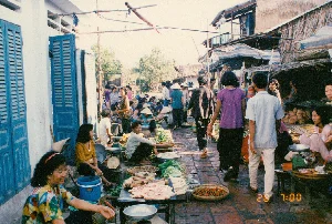 Cảnh một khu chợ quê ngoài trời ở Sóc Trăng vào buổi sáng sớm, khoảng 7 giờ ngày 28 tháng 4 năm 1994. Lối đi trong chợ được lát gạch đỏ, không khí mua bán rất tấp nập với nhiều người qua lại.。Những người bán hàng, phần lớn là phụ nữ, ngồi trên ghế đẩu thấp hoặc trực tiếp trên nền đất để bày bán hàng hóa. Một số phụ nữ đội nón lá. Các mặt hàng được bày bán rất phong phú, bao gồm thịt heo đã thái sẵn đặt trên bàn, các loại rau củ như củ cải trắng và rau xanh, cùng một mẹt lớn đầy ớt đỏ và xanh. Ở phía xa hơn, có thể thấy một sạp hàng bán dừa.。Nhiều chiếc cân đĩa (cân đồng hồ) được đặt cạnh hàng hóa để phục vụ việc mua bán. Mọi người sử dụng các loại rổ, rá, mẹt đan bằng tre để trưng bày sản phẩm. Bên trái là một ngôi nhà có tường sơn trắng và bộ cửa gỗ lá sách màu xanh lam. Bên phải là các gian hàng có kết cấu đơn sơ, một số có mái lợp bằng lá.。（日本語訳）。1994年4月28日午前7時頃、ソクチャン省の田舎にある屋外市場の早朝の光景。市場の通路は赤レンガで舗装されており、多くの人々が行き交い、売買の雰囲気は非常に賑やかである。商人たち、そのほとんどは女性で、低いスツールに座るか、直接地面に座って商品を並べている。何人かの女性はノンラーをかぶっている。販売されている商品は非常に豊富で、テーブルの上に置かれたカット済みの豚肉、大根や青菜などの野菜類、そして赤唐辛子と青唐辛子でいっぱいの大きな平たいざるが含まれている。人々は商品を陳列するために、竹で編んだ様々な種類の籠、ざる、平たいざるを使用している。左側には、白い壁と青いルーバー式の木製ドアのある家がある。右側には、簡素な作りの屋台が並び、中には葉で葺いた屋根のものもある。