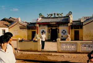 Bức ảnh chụp một ngôi chùa hoặc hội quán của người Hoa với kiến trúc đặc trưng, được sơn màu vàng. Trên nóc mái ngói là các chi tiết trang trí bằng gốm sứ, nổi bật là hình tượng 'lưỡng long chầu nguyệt' (hai con rồng chầu vào một vòng tròn ở giữa). Lối vào sân chùa được canh giữ bởi hai tượng sư tử đá (lân đá). Phía trước chùa là một con đường đất. Có một số người trong ảnh: một người đàn ông mặc áo thun trắng đang đi từ trong sân ra, một người khác đang ngồi nghỉ trong sân, và ở tiền cảnh là một vài thanh thiếu niên đang đứng nhìn. Trên các bức tường có nhiều biển hiệu ghi chữ Hán và chữ Quốc ngữ. Phía trên cửa chính có các biển hiệu lớn ghi '聖聖王三' (Thánh Thánh Vương Tam) và '運廣德帝' (Vận Quảng Đức Đế). Ở dãy nhà bên trái, có một tấm biển ghi 'LỚP HOA VĂN VĨNH PHƯỚC', và các dòng chữ Hán như '育教心熱' (Dục Giáo Tâm Nhiệt - Hết lòng với giáo dục). Ở dãy nhà bên phải, một tấm biển khác ghi '校學...' (Hiệu Học...), cho thấy nơi đây có thể vừa là nơi thờ tự, vừa là một trung tâm văn hóa hoặc trường học của cộng đồng. Xung quanh là các ngôi nhà lợp ngói và có thể thấy dây điện, ăng-ten TV, phù hợp với bối cảnh năm 1994.。（日本語訳）。写真は、黄色に塗られた特徴的な建築様式を持つ、華人の寺院または会館を撮影したものです。瓦屋根の上には陶磁器の装飾が施されており、特に「双龍朝月」（二匹の龍が中央の円に向かい合う）の像が目立ちます。寺院の境内への入り口は、二体の石獅子（麒麟の石像）によって守られています。寺院の前には未舗装の道があります。白いTシャツを着た男性が境内から出てくるところ、もう一人が境内で座って休んでいるところ、そして前景には数人の若者が立って見ています。壁には漢字とクオック・グー（ベトナム語のローマ字表記）で書かれた多くの看板があります。正面の扉の上には、「聖聖王三」（Thánh Thánh Vương Tam）と「運廣德帝」（Vận Quảng Đức Đế）と書かれた大きな看板があります。左側の建物には、「LỚP HOA VĂN VĨNH PHƯỚC」と書かれた看板があり、「育教心熱」（Dục Giáo Tâm Nhiệt - 教育に熱心）のような漢字の文字列もあります。右側の建物には、別の看板に「校學...」（Hiệu Học...）と書かれており、この場所が礼拝の場であると同時に、コミュニティの文化センターまたは学校でもある可能性を示しています。周囲には瓦葺きの家々があり、電線やテレビアンテナが見え、1994年という時代背景と一致しています。