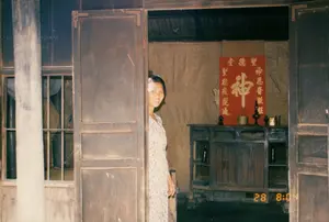 写真には、おそらくソクチャン省のある農村地域にある木造家屋の戸口に立っている一人の女性が写っている。その女性は花柄の服を着て、木製のドアの後ろから身をのぞかせ、撮影者に向かって微笑んでいる。祭壇の後ろの壁には、黄色い漢字が書かれた大きな赤い対聯が掛けられている。中央の最も大きな文字は「神」（Thần）である。両側の二つの縦書きの行は、「聖恩普照旺」（Thánh Ân Phổ Chiếu Vượng）と「聖德庇運通」（Thánh Đức Tí Vận Thông）である。祭壇の上には、線香が立てられた香炉、燭台、その他の祭具がある。家の建築はかなりシンプルで、軒先に大きな木の柱があり、出入り口のドアと窓は木製である。窓には鉄格子がある。外側の木の壁には手書きの文字の跡があるが、読むことはできない。写真の右下隅には、「28 8:04」と表示された黄色いデジタルのタイムスタンプがある