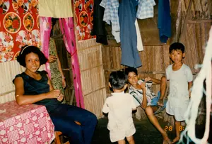 Bối cảnh là bên trong một ngôi nhà có kết cấu đơn sơ ở vùng nông thôn, có thể là ở khu vực Sóc Trăng vào năm 1994. Các bức tường của ngôi nhà được làm bằng vật liệu tự nhiên như lá hoặc tre đan, một kiểu kiến trúc nhà ở phổ biến ở Đồng bằng sông Cửu Long thời kỳ đó.。Hoạt động chính là một buổi sinh hoạt gia đình hoặc một khoảnh khắc thư giãn. Một phụ nữ trẻ đang ngồi bên chiếc bàn nhỏ, mỉm cười nhìn vào máy ảnh. Phía sau cô là ba cậu bé ở các độ tuổi khác nhau. Hai cậu bé lớn hơn đang ngồi ở góc phòng, trong khi một cậu bé nhỏ tuổi nhất đang đứng ở phía trước. Không khí chung toát lên vẻ đời thường và ấm cúng.。Các vật thể có trong ảnh bao gồm:。- Một chiếc bàn nhỏ được phủ khăn trải bàn hoa văn ca-rô đỏ trắng.。- Quần áo (áo sơ mi kẻ sọc, quần dài) được treo trên tường, cho thấy không gian sống đơn giản.。- Tường được trang trí bằng giấy dán nhiều màu sắc. Một loại giấy dán màu đỏ có họa tiết hoa hồng và chữ "Joy" có thể được nhìn thấy. Ngoài ra còn có các dải nhựa màu hồng được treo làm rèm cửa hoặc để trang trí.。- Trên tường có treo một tấm vải hoặc bao tải có in chữ nước ngoài (giống chữ Cyrillic) nhưng không thể đọc rõ.。（日本語訳）。背景は、1994年のソクチャン地方と思われる田舎の、簡素な造りの家の中です。家の壁は、葉や編んだ竹などの自然素材でできており、当時のメコンデルタで一般的だった住宅建築様式です。主な活動は、家族の集まりか、リラックスしたひとときです。若い女性が小さなテーブルのそばに座り、カメラに向かって微笑んでいます。彼女の後ろには、年齢の異なる3人の男の子がいます。年上の2人の男の子は部屋の隅に座っており、一番年下の男の子は手前に立っています。全体の雰囲気は、日常的で温かい様子を醸し出しています。写真に写っている物には以下が含まれます：。- 赤と白のチェック柄のテーブルクロスがかけられた小さなテーブル。- 壁に掛けられた衣服（ストライプのシャツ、ズボン）は、簡素な生活空間を示しています。- 壁は色とりどりの紙で飾られています。バラの模様と「Joy」という文字が入った赤い紙が見えます。また、カーテンや装飾としてピンク色のプラスチックの帯が吊るされています。- 壁には、外国の文字（キリル文字に似ている）が印刷された布か袋が掛けられていますが、はっきりと読むことはできません。