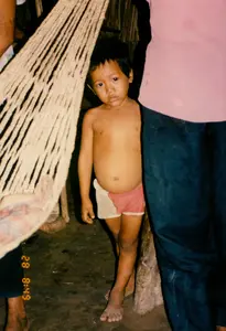 Trên ảnh có dòng chữ số màu vàng: '28 8:49'. Bức ảnh chụp cận cảnh một bé trai đang đứng bên trong một ngôi nhà đơn sơ ở vùng nông thôn. Cậu bé ở trần, mặc một chiếc quần đùi hai màu hồng và trắng, đi chân đất trên nền đất. Cậu bé có vẻ mặt trầm ngâm, nhìn thẳng vào ống kính. Bên cạnh cậu bé, phía bên trái là một chiếc võng lưới được đan bằng sợi thô, màu trắng ngà. Phía sau và xung quanh cậu bé là những người lớn, nhưng chỉ nhìn thấy một phần cơ thể của họ: một người mặc áo hồng và quần sẫm màu đứng bên phải, và một phần chân của người khác ở bên trái. Khung cảnh cho thấy một không gian sống đơn giản với nền đất và cột nhà bằng gỗ thô sơ.。（日本語訳）。写真には黄色い数字の列「28 8:49」があります。写真は、田舎の質素な家の中に立っている男の子のクローズアップです。男の子は上半身裸で、ピンクと白の2色の半ズボンを履き、土の床の上で裸足です。男の子は物思いにふけるような表情で、カメラのレンズをまっすぐ見ています。男の子の隣、左側には、粗いオフホワイトの糸で編まれた網のハンモックがあります。男の子の後ろや周りには大人がいますが、彼らの体の一部しか見えません。右側に立っているピンクのシャツと暗い色のズボンを履いた人、そして左側には別の人間の脚の一部が見えます。その光景は、土の床と粗末な木製の家の柱がある、シンプルな生活空間を示しています。