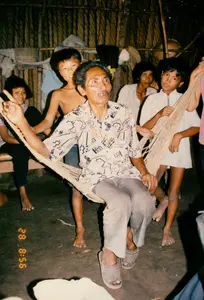 この写真は、農村地帯にある葉葺きの家の中での日常的な生活の一場面を捉えたもので、1994年にソクチャンで撮影された可能性があります。家は簡素な造りで、壁は葉で編まれ、床は土間です。中心人物は、粗い繊維で作られた網のハンモックに座ってくつろいでいる年配の男性です。彼は柄物のシャツと長ズボンを着用し、プラスチックのサンダルを履いています。彼は楽しそうに話しているか歌っており、同時に手を挙げてVサインをしています。彼の周りには子供たちのグループがおり、おそらく家族の子供や孫、あるいは近所の子たちでしょう。何人かの子供は裸足で、一人の男の子は上半身裸で彼の後ろに立ち、別の男の子はハンモックにつかまっています。写真の雰囲気は、当時のベトナムの田舎における家族生活の温かさ、素朴さ、そして親密さを醸し出しています。後方には、竹の竿に掛けられた衣類が見えます。写真の左下隅には、「28 8:56」と記されたオレンジ色のデジタルタイムスタンプがあります