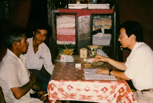 Chữ viết: Trên chiếc mũ lưỡi trai màu trắng đặt trên bàn có chữ "AHA" màu đỏ, có thể là một phần của chữ "YAMAHA". Trên một tấm vải màu hồng bên trong tủ kính phía sau có chữ viết nhưng không thể đọc rõ. Dấu ngày tháng màu vàng ở góc dưới bên phải của bức ảnh ghi "94 4 28".。Bối cảnh và hoạt động: Bối cảnh là bên trong một ngôi nhà, có thể là phòng khách. Ba người đàn ông đang ngồi quanh một chiếc bàn. Người đàn ông bên phải, đeo kính và mặc áo sơ mi trắng, dường như đang tiến hành một cuộc phỏng vấn hoặc thảo luận, tay chỉ vào giấy tờ đặt trên bàn. Hai người đàn ông còn lại, một người lớn tuổi và một người trẻ hơn, đang lắng nghe một cách chăm chú. Người đàn ông lớn tuổi đang cầm một điếu thuốc lá. Hoạt động này trông giống như một cuộc khảo sát, phỏng vấn hoặc một buổi làm việc tại nhà dân.。Các vật thể được nhìn thấy: Ba người đàn ông, một chiếc bàn gỗ được phủ khăn trải bàn bằng ni lông có hoa văn màu đỏ và trắng. Trên bàn có một chiếc mũ lưỡi trai, một nải chuối, một bình trà bằng gốm, một cái bát trang trí, một hộp gỗ nhỏ (có thể là hộp đựng con dấu), và các loại giấy tờ. Phía sau họ là một chiếc tủ gỗ có cửa kính, bên trong có chứa vải vóc hoặc quần áo gấp lại. Một bóng đèn tuýp được lắp đặt phía trên tủ.。（日本語訳）。文字：テーブルの上に置かれた白い野球帽には赤い文字で「AHA」と書かれており、それは「YAMAHA」という文字の一部である可能性があります。写真の右下隅にある黄色い日付スタンプには「94 4 28」と記されています。背景と活動：背景は家の中、おそらくリビングルームです。3人の男性がテーブルを囲んで座っています。右側の男性は、眼鏡をかけ白いシャツを着ており、インタビューか議論を行っているようで、テーブルの上に置かれた書類を指さしています。残りの2人の男性、1人は年配で、もう1人は若く、熱心に耳を傾けています。年配の男性はタバコを手に持っています。この活動は、調査、インタビュー、または民家での作業セッションのように見えます。テーブルの上には、野球帽1つ、バナナ一房、陶器のティーポット1つ、装飾的なボウル1つ、小さな木箱（印鑑ケースの可能性があります）、そして各種書類があります。彼らの後ろにはガラス戸の付いた木製の戸棚があり、中には布地や折りたたまれた衣類が入っています。戸棚の上には蛍光灯が設置されています。