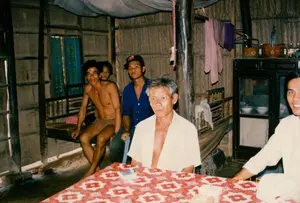 Bối cảnh là bên trong một ngôi nhà ở vùng nông thôn, có kết cấu đơn sơ với vách làm bằng vật liệu tự nhiên như tre, lá, và nền đất. Một nhóm người, chủ yếu là đàn ông, đang tụ tập trong nhà. Ở phía trước, một người đàn ông lớn tuổi mặc áo sơ mi trắng không cài cúc ngồi bên một chiếc bàn được trải khăn hoa văn màu đỏ và trắng. Ở phía sau, một người đàn ông cởi trần ngồi trên một chiếc phản gỗ, bên cạnh là một thanh niên mặc áo sơ mi xanh và đội mũ lưỡi trai. Một người phụ nữ cũng có mặt ở phía sau. Hoạt động chính là một buổi tụ họp, trò chuyện thân mật. Các đồ vật trong nhà bao gồm một chiếc tủ gỗ có cửa kính đựng chén bát, một chiếc võng được mắc lên, và quần áo đang được phơi. Có một cửa sổ chớp bằng gỗ màu xanh lam ở phía sau. Trên bàn có một bao thuốc lá và tiền giấy. Không có văn bản hay chữ viết nào có thể đọc được trong ảnh.。（日本語訳）。背景は田舎のある家の中、竹や葉のような自然素材で作られた壁と土間を持つ素朴な造りである。主に男性からなる一団が家の中に集まっている。手前では、ボタンを留めていない白いシャツを着た年配の男性が、赤と白の模様のテーブルクロスが敷かれたテーブルのそばに座っている。後ろでは、上半身裸の男性が木製の寝台に座っており、その隣には青いシャツを着て野球帽をかぶった若者がいる。後ろには女性も一人いる。主な活動は、親密な会話を交わす集まりである。家の中の物には、食器が入ったガラス戸の木製戸棚、吊るされたハンモック、そして干されている衣類が含まれる。後ろには青い木製のよろい戸の窓がある。テーブルの上にはタバコの箱と紙幣がある。写真の中には、読み取れるテキストや文字はない。