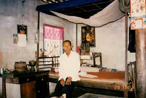 Bối cảnh là bên trong một ngôi nhà ở vùng nông thôn, có thể là ở Sóc Trăng. Một người đàn ông lớn tuổi mặc áo sơ mi trắng và quần sẫm màu đang ngồi trên mép một chiếc giường gỗ. Chiếc giường có khung và một chiếc mùng (màn) màu trắng được treo phía trên. Trên giường có trải chiếu và gối. Phía sau người đàn ông là một cửa sổ có rèm che và một bàn thờ nhỏ. Trên bàn thờ có một bức tranh thờ Phật, chân nến và nhang. Bên trái có một chiếc bàn gỗ, trên đó đặt một cái giỏ tre, một cái thố bằng đồng và vài vật dụng khác. Trên bức tường vôi đã cũ, có treo một tờ lịch bloc hiển thị rõ ngày 28, một bức ảnh của một bé gái và một tờ quảng cáo nhỏ nhưng không rõ nội dung. Một chiếc mũ được treo trên cột nhà. Người đàn ông đang nhìn thẳng vào máy ảnh, dường như đang được chụp ảnh. Không gian trong phòng khá đơn giản và mộc mạc, phản ánh cuộc sống ở nông thôn Việt Nam vào năm 1994.。（日本語訳）。背景は田舎の家の中、おそらくソクチャン省だろう。白いシャツと濃い色のズボンを履いた年配の男性が、木製のベッドの端に座っている。ベッドにはフレームがあり、上には白い蚊帳が吊るされている。ベッドの上にはござと枕が敷かれている。男性の後ろにはカーテンのかかった窓と小さな祭壇がある。祭壇の上には仏様の絵、燭台、線香がある。古くなった漆喰の壁には、28日とはっきり表示された日めくりカレンダー、女の子の写真、そして内容は不明だが小さな広告が掛けられている。家の柱には帽子が掛けられている。男性はカメラをまっすぐ見ており、写真を撮られているようだ。部屋の空間は非常にシンプルで素朴であり、1994年当時のベトナムの田舎の生活を反映している。