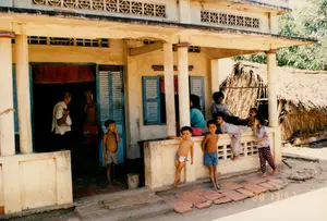 Bối cảnh là một ngôi nhà ở vùng nông thôn, có thể là ở Sóc Trăng, vào một buổi chiều. Hình ảnh cho thấy một ngôi nhà chính một tầng, xây bằng bê tông, có hiên và các cột tròn. Bên cạnh là một ngôi nhà phụ có kết cấu đơn giản hơn với mái lợp bằng lá. Nhiều người, chủ yếu là trẻ em và một vài người lớn, đang tụ tập trước và trong nhà. Trên hiên nhà, một vài phụ nữ và trẻ em đang ngồi và nằm nghỉ. Các bé gái mặc đồ bộ, trong khi các bé trai thì cởi trần, chỉ mặc quần đùi. Hai cậu bé đang đứng chân trần trên nền gạch tàu đã vỡ phía trước hiên. Một cậu bé khác đứng ở ngưỡng cửa. Bên trong cửa chính, có thể thấy một người đàn ông lớn tuổi đầu trọc, mặc áo sơ mi trắng, và một người đàn ông khác cởi trần. Ngôi nhà bê tông có các chi tiết kiến trúc đặc trưng của thời kỳ đó như cửa sổ lá sách màu xanh lam và gạch bông gió để thông gió. Một tấm rèm màu đỏ được treo ở cửa. Phía trên một cánh cửa có một tấm biển nhưng chữ viết trên đó không thể đọc được. Ở góc dưới bên phải của bức ảnh có dấu ngày tháng và thời gian là '28 13:57'.。（日本語訳）。背景は、ある日の午後、田舎の一軒の家で、おそらくソクチャン省にあると思われます。写真には、コンクリート造りの一階建ての母屋が写っており、軒先と丸い柱があります。その隣には、葉で葺いた屋根を持つ、より簡素な造りの離れがあります。主に子供たちと数人の大人からなる多くの人々が、家の前と中に集まっています。軒先では、数人の女性と子供たちが座ったり横になったりして休んでいます。女の子たちはセットアップの服を着ていますが、一方、男の子たちは上半身裸で、短パンだけを履いています。軒先の前の割れたテラコッタタイルの床の上に、二人の男の子が裸足で立っています。別の男の子が戸口に立っています。正面の戸口の中には、白いシャツを着た禿頭の年配の男性と、上半身裸の別の男性が見えます。コンクリートの家には、青いルーバー窓や換気のための透かしブロックなど、その時代の特徴的な建築様式の細部が見られます。赤いカーテンが戸口に掛けられています。ある扉の上には看板がありますが、そこに書かれている文字は読み取れません。写真の右下隅には、「28 13:57」という日付と時刻のスタンプがあります。