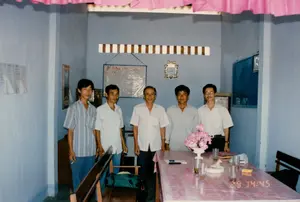 Bối cảnh là một căn phòng trông giống như một phòng họp hoặc văn phòng tại Sóc Trăng vào năm 1994. Năm người đàn ông Việt Nam đang đứng chụp ảnh kỷ niệm phía sau một chiếc bàn dài. Chiếc bàn được phủ một tấm khăn trải bàn bằng nhựa màu hồng, trên đó có đặt một lọ hoa giả màu hồng, vài chiếc cốc thủy tinh, một cuốn sổ tay và một bức tượng nhỏ. Những người đàn ông mặc áo sơ mi và quần dài, một người đang cầm điếu thuốc lá. Xung quanh có ghế gỗ và một chiếc túi màu xanh lá cây. Các bức tường được sơn màu xanh nhạt. Trên tường phía sau có treo một chiếc đồng hồ, một vật trang trí sọc và một tấm bảng trắng có chữ viết nhưng không thể đọc được. Trên tường bên trái có ghim một số tài liệu. Trần nhà được trang trí bằng các dải diềm xếp nếp màu hồng. Trên ảnh có dấu thời gian kỹ thuật số ghi '29 14:45'.。（日本語訳）。舞台は1994年のソクチャンにある、会議室かオフィスのような部屋です。5人のベトナム人男性が、長いテーブルの後ろに立って記念撮影をしています。テーブルにはピンク色のビニール製テーブルクロスが敷かれ、その上にはピンク色の造花の花瓶、いくつかのガラスのコップ、一冊の手帳、そして小さな置物が置かれています。男性たちはシャツと長ズボンを着用しており、一人はタバコを手に持っています。周りには木製の椅子と緑色のバッグがあります。壁は水色に塗られています。後ろの壁には、時計、ストライプ柄の装飾品、そして文字が書かれているが読めないホワイトボードが掛けられています。左側の壁には、いくつかの書類がピンで留められています。天井はピンク色のフリルのついた飾り帯で装飾されています。写真には「29 14:45」と記されたデジタルのタイムスタンプがあります。