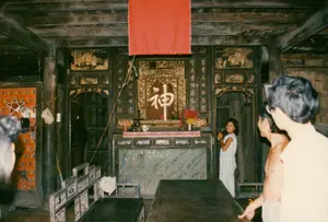 Bối cảnh là không gian bên trong một ngôi nhà gỗ cổ hoặc một gian thờ tự, có thể là nhà của một gia đình người Hoa ở Sóc Trăng. Kiến trúc đặc trưng với cột và xà nhà bằng gỗ sẫm màu. Tâm điểm của căn phòng là một bàn thờ tổ tiên lớn, được chạm khắc tinh xảo, sơn son thếp vàng. Ở chính giữa bàn thờ có một bài vị lớn với chữ Hán "神" (Thần) màu trắng trên nền đỏ. Phía trên bàn thờ có treo một tấm vải đỏ. Trên bàn thờ bày các vật phẩm cúng kiếng như ly, chén và chân nhang. Hai bên bàn thờ có hai cặp câu đối chữ Hán được khắc trên các tấm gỗ. Các chữ có thể nhận diện là: Vế phải (nhìn từ ngoài vào): "富貴花開金盞" (Phú quý hoa khai kim trản) và "寶馬香烟騰紫氣" (Bảo mã hương yên đằng tử khí). Vế trái (nhìn từ ngoài vào): "銀燭全堂耀祥光" (Ngân chúc toàn đường diệu tường quang) và "平安二字值千金" (Bình an nhị tự trị thiên kim). Trong phòng có một số người đang đứng quan sát. Một cô bé mặc váy hoa đang đứng gần bàn thờ và nhìn về phía máy ảnh. Ngoài ra còn có một người đàn ông cởi trần và một vài người khác. Các đồ vật khác trong phòng bao gồm một bộ bàn ghế gỗ kiểu cổ ở bên trái, một chiếc bàn gỗ lớn ở phía trước. Một chiếc nón lá được treo trên cột nhà. Trên bức tường phía xa bên trái được dán giấy hoa văn màu cam, có một ngôi sao năm cánh màu trắng và vài tấm ảnh nhỏ được ghim lên.。（日本語訳）。背景は、古い木造家屋または祭祀空間の内部で、ソクチャン省の華人家庭の家である可能性があります。建築は、濃い色の木製の柱と梁が特徴です。部屋の中心には、精巧に彫刻され、朱塗り金箔で装飾された大きな先祖代々の祭壇があります。祭壇の中央には、赤い背景に白い漢字「神」（Thần）が書かれた大きな位牌があります。祭壇の上には赤い布が掛けられています。祭壇の上には、杯、碗、香炉などの供物が置かれています。祭壇の両側には、木の板に刻まれた漢字の対聯が二対あります。識別できる文字は次のとおりです。右側（外から見て）：「富貴花開金盞」（Phú quý hoa khai kim trản）と「寶馬香烟騰紫氣」（Bảo mã hương yên đằng tử khí）。左側（外から見て）：「銀燭全堂耀祥光」（Ngân chúc toàn đường diệu tường quang）と「平安二字值千金」（Bình an nhị tự trị thiên kim）。部屋の中には数人が立って見物しています。花柄のスカートをはいた少女が祭壇の近くに立ち、カメラの方を見ています。その他に、上半身裸の男性と他の数人がいます。部屋にある他の物には、左側に古い様式の木製テーブルと椅子のセット、手前に大きな木製のテーブルが含まれます。ノンラー（ベトナムの伝統的な笠）が家の柱に掛けられています。左奥の壁にはオレンジ色の模様の紙が貼られており、白い五芒星と数枚の小さな写真がピンで留められています。