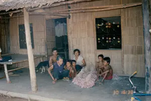 Bức ảnh chụp cảnh một gia đình nhiều thế hệ đang quây quần trước hiên một ngôi nhà ở vùng nông thôn. Ngôi nhà có kiến trúc đơn sơ, đặc trưng của vùng quê Việt Nam thời bấy giờ, với vách được làm bằng vật liệu đan lát như tre hoặc lá dừa nước, mái lợp lá, và nền đất. Bên trong hiên nhà, có một cái chõng tre.。Gia đình gồm một bà cụ, một phụ nữ, một thanh niên và bốn đứa trẻ đang ngồi trên nền đất. Ở giữa họ là một đống hành tím rất lớn đã được thu hoạch và bó lại thành từng chùm. Hoạt động này cho thấy họ có thể đang trong quá trình sơ chế nông sản sau thu hoạch, có thể là để bán hoặc dự trữ.。Các vật thể khác trong ảnh bao gồm một con chó đang đi lại gần đó, một phần của chiếc xe đạp ở góc phải, và quần áo phơi bên trong cửa ra vào. Cửa sổ của ngôi nhà có song sắt và được trang trí bằng những bông hoa nhựa nhiều màu sắc. Ở góc dưới bên phải bức ảnh có dấu thời gian màu cam ghi '30 16:38'.。（日本語訳）。田舎のある家の軒先で、多世代の家族が集まっている光景を写した写真。その家は、壁は竹やニッパヤシの葉のような編んだ素材で作られ、屋根は葉で葺かれ、そして土間という、当時のベトナムの田舎に特徴的な素朴な建築様式である。軒の内側には、竹製の寝台が一つある。家族は、一人のおばあさん、一人の女性、一人の青年、そして4人の子供からなり、土間に座っている。彼らの真ん中には、収穫されて束にまとめられた赤玉ねぎの大きな山がある。この活動は、彼らが収穫後の農産物を一次加工している過程にある可能性を示しており、それは販売または貯蔵のためかもしれない。写真の中の他の物には、近くを歩き回っている一匹の犬、右隅にある自転車の一部、そして出入り口の内側に干されている服が含まれる。家の窓には鉄格子があり、色とりどりのプラスチックの花で飾られている。写真の右下隅には、「30 16:38」と記されたオレンジ色のタイムスタンプがある。