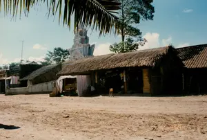 Bức ảnh chụp một khung cảnh ở vùng nông thôn, có khả năng là ở Sóc Trăng, vào một ngày nắng. Mặt đất là một khoảng sân rộng bằng đất cát, không được lát đá.。Ở trung tâm là một dãy nhà lợp mái lá, trông giống như một cửa hàng tạp hóa hoặc một quán nhỏ. Phía trước cửa hàng có một quầy hàng tạm được che bằng tấm bạt sọc xanh, đỏ và trắng. Bên trong cửa hàng tối, có thể thấy một vài người và một số hàng hóa. Một chiếc ghế đẩu gỗ được đặt ở ngoài sân. Có một tấm biển dọc màu trắng treo ở cửa hàng nhưng chữ viết trên đó không thể đọc được.。Ở phía sau những ngôi nhà mái lá, có thể nhìn thấy một công trình kiến trúc nổi bật, dường như là cổng của một ngôi chùa Khmer với nhiều tầng mái và các chi tiết trang trí phức tạp, một đặc điểm kiến trúc phổ biến ở Sóc Trăng.。Bên trái, có một bức tường thấp bằng xi măng với các cột trang trí có tượng nhỏ hình sư tử trên đỉnh. Một chiếc ăng-ten TV có thể được nhìn thấy trên một mái nhà ở phía xa. Phía trên cùng bên trái của bức ảnh là một tàu lá dừa hoặc lá cọ. Hoạt động chính dường như là sinh hoạt buôn bán thường ngày tại một khu dân cư gần một ngôi chùa.。（日本語訳）。写真は、晴れた日の、おそらくソクチャン省の田舎の風景を写したものです。地面は舗装されていない砂地の広い庭です。店の前には、青、赤、白の縞模様の防水シートで覆われた仮設の屋台があります。店内は暗く、数人の人々といくつかの商品が見えます。木製の丸椅子が庭に置かれています。店には縦長の白い看板が掛かっていますが、そこに書かれている文字は読めません。茅葺き屋根の家々の後ろには、ソクチャン省でよく見られる建築様式である、幾層にも重なった屋根と複雑な装飾が施されたクメール寺院の門と思われる、際立った建築物が見えます。左側には、上に小さなライオンの像が乗った装飾的な柱のある、セメント製の低い壁があります。遠くの屋根の上にはテレビアンテナが見えます。写真の左上には、ココナッツかヤシの葉が見えます。主な活動は、寺院の近くの住宅地での日常的な商売のようです。