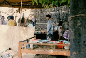 Bối cảnh là một quán ăn đơn sơ, có thể ở vùng nông thôn hoặc một khu chợ địa phương vào khoảng năm 1994. Quán được dựng tạm bằng các vật liệu tự nhiên, với mái lợp lá và vách làm bằng lá dừa nước khô đan lại, được chống đỡ bằng các cột tre. Nền đất. Ở phía trước, một chiếc giường gỗ (hoặc chõng tre) được dùng làm nơi để đồ, trên đó có nhiều thau nhựa lớn màu đỏ và xanh, mâm nhôm và các chồng chén bát. Bên trong quán, một người đàn ông mặc áo sơ mi hoa văn đang đi qua. Phía sau anh là một bé gái. Trên quầy hàng phía sau, nhiều chén nhỏ đựng thức ăn được bày sẵn, bên cạnh có một chiếc nồi nhôm lớn. Ở hậu cảnh bên trái, một nhóm phụ nữ đang đứng nói chuyện với nhau. Khung cảnh cho thấy một hoạt động buôn bán đồ ăn thức uống thường ngày, phản ánh đời sống sinh hoạt giản dị ở miền quê Việt Nam vào thời điểm đó. Trên hình có dấu thời gian "2 7:51".。（日本語訳）。背景は、1994年頃の田舎か地元の市場にある質素な食堂です。食堂は、葉で葺かれた屋根と、乾燥したニッパヤシの葉を編んで作られた壁を持ち、竹の柱で支えられた、自然素材で仮設されたものです。地面は土です。正面には、木製のベッド（または竹製の寝台）が物を置く場所として使われており、その上には赤と青の大きなプラスチック製のたらい、アルミ製の盆、そして積み重ねられた食器類がたくさんあります。店内では、柄物のシャツを着た男性が通り過ぎています。彼の後ろには一人の女の子がいます。後方のカウンターには、食べ物が入った多くの小鉢が並べられており、その隣には大きなアルミ製の鍋があります。左の背景では、女性たちのグループが立ち話をしています。この光景は、日常的な飲食物の商売活動を示しており、当時のベトナムの田舎における質素な生活を反映しています。画像には「2 7:51」というタイムスタンプがあります。