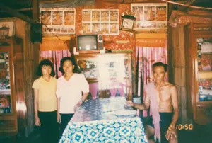 Bức ảnh chụp cảnh sinh hoạt bên trong một ngôi nhà ở Sóc Trăng vào năm 1994. Đây có thể là phòng khách của một gia đình. Có ba người trong ảnh: một người đàn ông lớn tuổi đang ngồi bên bàn, ông cởi trần, mặc quần đùi và vắt một chiếc khăn rằn (krama) trên vai. Đứng bên cạnh là một phụ nữ trung niên mặc áo hồng và một cô gái trẻ mặc áo vàng. Họ đang cùng nhau chụp ảnh kỷ niệm. Căn phòng có vách làm bằng vật liệu đan lát như tre, nứa. Ở giữa là một chiếc bàn được phủ khăn trải bàn hoa văn màu xanh và trắng. Phía sau có một chiếc tủ gỗ lớn có cửa kính, trên nóc tủ đặt một chiếc tivi kiểu cũ. Các bức tường được trang trí bằng nhiều tấm lịch hoặc tranh ảnh có hình ảnh Phật giáo (có thể là Phật giáo Nam tông Khmer), nhưng chữ viết trên đó không thể đọc được. Ngoài ra còn có đồng hồ treo tường, một chiếc loa và các tấm vải nhiều màu sắc dùng làm rèm cửa. Dấu ngày tháng của máy ảnh hiển thị "2 10:50" ở góc dưới bên phải.。（日本語訳）。1994年にソクチャン省のある家の中での生活風景を撮影した写真です。これは、ある家族の居間だと思われます。年配の男性がテーブルのそばに座っており、上半身は裸で、半ズボンを履き、肩にはクラマー（krama）を掛けています。その隣には、ピンクの服を着た中年の女性と、黄色の服を着た若い女の子が立っています。彼らは一緒に記念写真を撮っています。部屋の壁は、竹やニッパヤシのような編んだ素材でできています。中央には、青と白の模様のテーブルクロスで覆われたテーブルがあります。後ろにはガラス戸の付いた大きな木製の棚があり、棚の上には旧式のテレビが置かれています。壁は、仏教（おそらくクメールの上座部仏教）の絵が描かれた多くのカレンダーや絵で飾られていますが、そこに書かれている文字は読むことができません。その他にも、壁掛け時計、スピーカー、そしてカーテンとして使われている色とりどりの布があります。カメラの日付スタンプは、右下に「2 10:50」と表示されています。