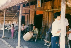 背景は農村部にある高床式の家の軒先で、おそらくソクチャン省だと思われる。家は木造で、屋根は葉（おそらくニッパヤシの葉）で葺かれており、軒下の床はタイル張りである。出入り口の上部の木の壁には、小さなカラーの絵が掛けられており、それは動物に乗る神が描かれたクメール人の祭壇画のようである。木の壁にはいくつかの数字と文字が書かれているが、はっきりと読み取ることはできない。活動については、一人の男性が木の椅子に座って手作業をしており、具体的には竹や笹で大きなカゴか魚を捕る罠を編んでいる。彼の隣には、赤ちゃんを抱いた女性が座っている。庭の方では、ピンク色の服を着た女の子が立って見ている。右側には、一団の人々が立って話している。注目すべき物としては、よろい戸の窓がある木造の家、木の柱、木の背もたれ椅子、後ろにある手押し車、そしてタイル張りの床に置かれたほうきなどが挙げられる