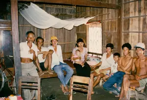 Trên vách tường gỗ có ghi các con số viết tay: "46-5" và "35-2".。Bối cảnh là bên trong một ngôi nhà ở nông thôn, có kết cấu đơn giản với khung và cột bằng gỗ, vách tường bằng gỗ và phên tre/lá. Một nhóm người, dường như là một gia đình nhiều thế hệ hoặc hàng xóm, đang tụ tập và chụp ảnh. Nhóm người này bao gồm nhiều người đàn ông ở các độ tuổi khác nhau, một phụ nữ đang bế một em bé sơ sinh, và một bé trai. Mọi người đang ngồi trên một chiếc phản gỗ dài và vài chiếc ghế gỗ đơn sơ. Một số người đàn ông đang cầm thuốc lá. Phía trên đầu họ có một chiếc mùng (màn) màu trắng được treo lên xà nhà. Ở góc trái có một chiếc xe đạp. Không khí chung cho thấy một buổi sum họp gia đình hoặc gặp gỡ thân mật, phản ánh cuộc sống sinh hoạt giản dị ở vùng nông thôn Việt Nam thời kỳ đó.。（日本語訳）。木の壁には「46-5」と「35-2」という手書きの数字が書かれている。背景は田舎の家の中であり、木製の骨組みと柱、木製の壁、竹や葉で編んだ壁というシンプルな構造である。多世代家族か隣人と思われる人々のグループが集まって写真を撮っている。このグループには、さまざまな年齢の男性数人、新生児を抱いた女性一人、そして男の子一人が含まれている。人々は長い木製の寝台といくつかの簡素な木製の椅子に座っている。何人かの男性はタバコを持っている。彼らの頭上には、梁から吊るされた白い蚊帳がある。左隅には自転車が一台ある。全体の雰囲気は、家族の集まりや親しい会合を示しており、当時のベトナムの田舎での質素な生活を反映している。