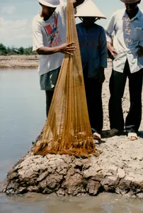 撮影背景は、農村地域にある水産養殖場で、エビまたは魚の養殖池である可能性があります。3人の男性が、乾いた土で盛られた池の岸に立っています。「CANADIAN」という文字が入った白いTシャツを着て、短パンを履き、帽子をかぶった若い男性が、両手で大きな黄色の漁網を持ち上げています。網の底の部分には、収穫されたばかりの小さな魚かエビが入っています。彼の隣には、ノンラー（菅笠）をかぶり、青いシャツと長ズボンを履いた年配の男性が立っています。3人目の男性は右側に立っており、明るい色のシャツと暗い色のズボンを履き、網の中を覗き込んで微笑んでいます。写真には「2 12:45 \'94」と記されたオレンジ色のデジタル日付スタンプがあり、写真が1994年5月2日に撮影されたことを示しています
