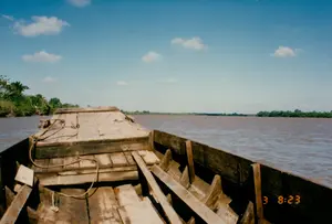 Bức ảnh được chụp từ góc nhìn trên một chiếc ghe gỗ đang di chuyển trên một con sông rộng, có khả năng là ở Sóc Trăng. Chiếc ghe gỗ cũ, được làm từ nhiều tấm ván, chiếm phần lớn tiền cảnh. Nước sông có màu nâu đục đặc trưng của phù sa. Hai bên bờ sông là những hàng cây xanh rậm rạp. Bầu trời trong xanh, có vài gợn mây trắng, cho thấy thời tiết đẹp. Hoạt động chính là di chuyển bằng ghe trên sông. Ở góc dưới bên phải của bức ảnh có dấu thời gian kỹ thuật số màu cam ghi '3 8:23', có thể là ngày 3 và thời gian là 8 giờ 23 phút sáng.。（日本語訳）。写真は、広い川の上を移動している一艘の木造の船の上からの視点で撮影されたもので、ソクチャン省である可能性があります。多くの板から作られた古い木造の船が、前景の大部分を占めています。川の水は、沖積土に特有の濁った茶色をしています。川の両岸は、緑豊かな木々の列です。空は青く澄み、白い雲がいくつかあり、天気が良いことを示しています。主な活動は、船で川の上を移動することです。写真の右下隅には、「3 8:23」と記されたオレンジ色のデジタルタイムスタンプがあり、3日の午前8時23分である可能性があります。