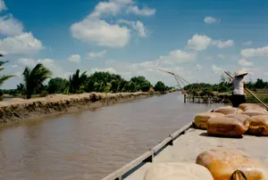 Bối cảnh là một con kênh ở vùng nông thôn, có khả năng là ở Sóc Trăng, dưới bầu trời xanh và có mây. Hai bên bờ kênh là bờ đất, được trồng nhiều cây dừa và các loại cây xanh khác. Nước kênh có màu nâu đục của phù sa. Trên mặt nước, một người đội nón lá đang đứng trên một chiếc ghe gỗ, dùng một cây sào dài để đẩy ghe di chuyển. Chiếc ghe đang chở rất nhiều can nhựa lớn màu vàng, có thể dùng để vận chuyển nước hoặc nhiên liệu. Ở phía xa dọc theo con kênh, có thể thấy những cái đó hoặc vó, là một ngư cụ đánh bắt cá truyền thống được làm bằng tre và lưới, dựng cố định trên mặt nước. Trên một trong những chiếc can nhựa có các ký tự mờ nhưng không thể đọc rõ.。（日本語訳）。背景は、おそらくソクチャン省の田舎にある運河で、空は青く雲がかかっています。運河の両岸は土手で、多くのココヤシの木やその他の緑の木々が植えられています。運河の水は、沖積土で濁った茶色をしています。水面では、ノンラーをかぶった人が木製の小舟の上に立ち、長い竿を使って舟を押し進めています。その小舟は、たくさんの大きな黄色のプラスチック製タンクを運んでおり、水や燃料を運ぶのに使われる可能性があります。運河沿いの遠くには、「đó」または「vó」が見えます。これは竹と網で作られた伝統的な漁具で、水面に固定して設置されています。プラスチック製タンクの一つには、ぼやけた文字がありますが、はっきりと読むことはできません。
